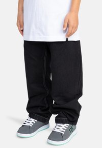 Jeans negros holgados con costuras blancas, combinados con zapatillas grises que tienen cordones blancos y una suela de color verde menta. Se puede ver una camiseta blanca.