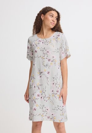 Femme aux cheveux bouclés portant une robe courte à manches courtes gris clair avec des motifs floraux roses et jaunes, debout devant un fond uni.