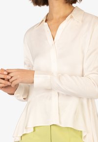 Blusa a maniche lunghe color crema con chiusura a bottoni frontale, orlo a peplum e texture liscia e lucida, abbinata a pantaloni verde chiaro.