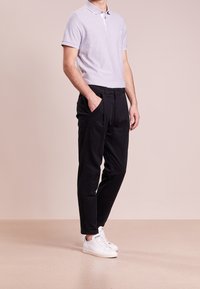 Helles lilafarbene Polohemd mit kurzen Ärmeln und weißem Kragen, kombiniert mit schwarzen Hosen und weißen Sneakers, vor einem neutralen Hintergrund stehend.