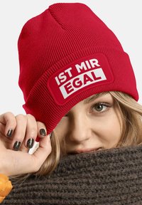 Red knit beanie with a rectangular patch featuring white text "IST MIR EGAL." The hat has ribbed texture and a snug fit. Hinted dark fur of a scarf below.