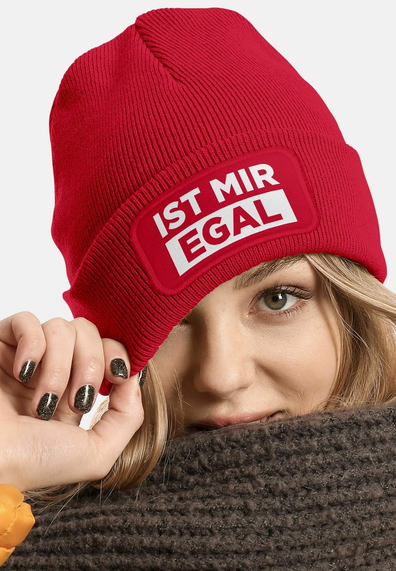Red knit beanie with a rectangular patch featuring white text "IST MIR EGAL." The hat has ribbed texture and a snug fit. Hinted dark fur of a scarf below.