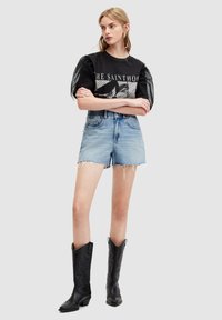 Camiseta gráfica negra y gris con mangas de malla abullonadas, combinada con shorts de mezclilla deshilachados. El modelo lleva botas vaqueras negras con punta afilada.