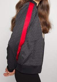 Svart bomberjacka med ett upprepande mönster, röda satinfickor på ärmarna, ribbade mudd och nederkant, dragkedjestängning och sidofickor.