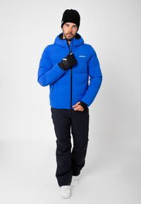 Veste isolante bleue avec capuche, fermeture éclair et détail du logo, associée à un pantalon noir et des gants, sur fond blanc.