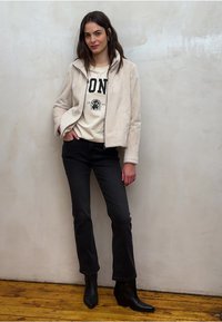 Crèmekleurig faux fur jack, witte trui met zwarte letters, zwarte uitlopende jeans en zwarte puntige enkellaarsjes; staand op houten vloer.