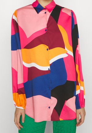 Langærmet skjorte med knapper og abstrakte, dristige former i pink, gul, blå, sort og hvid, båret med teksturerede grønne bukser.