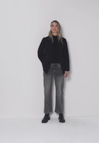 Kvinde iført sort blazer, sort top, grå vidbukser og sorte støvler, stående mod en ensfarvet lysegrå væg.