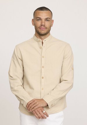 Chemise beige à manches longues avec col montant, boutons en bois et manches retroussables. Fabriquée en tissu doux avec une texture subtile.