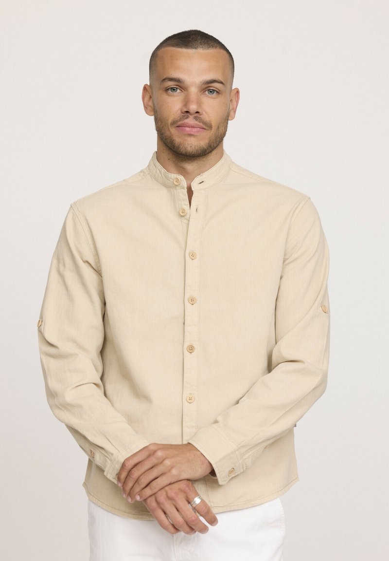 Chemise beige à manches longues avec col montant, boutons en bois et manches retroussables. Fabriquée en tissu doux avec une texture subtile.