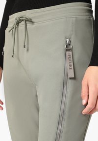 Ljust gröna byxor med en texturerad tygkvalitet, med en dragkedjeficka med en silverfärgad dragkedja och en grå etikett märkt "FUTURE" fäst.