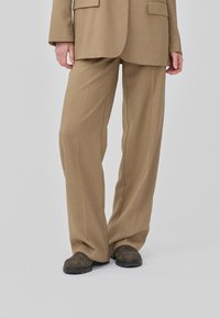 Pantalons taillés brun clair avec une coupe évasée, fabriqués en tissu texturé, associés à des chaussures sombres. Un look simple et structuré.
