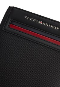 Μαύρο πορτοφόλι με υφή, με το λογότυπο Tommy Hilfiger και χαρακτηριστική λεπτομέρεια με ρίγα σε κόκκινο, λευκό και μπλε χρώμα.