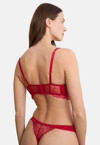 Frau trägt roten Spitzen-BH und passenden Tangaslip, von hinten vor einem schlichten weißen Hintergrund gezeigt.