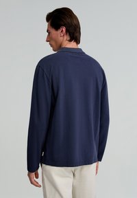 Polo shirt cu mânecă lungă de culoare bleumarin, cu un finisaj texturat și guler. Include slituri laterale și o etichetă mică a brandului la tiv.