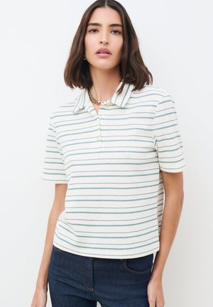 Nainen, jolla on olkapäille ulottuvat ruskeat hiukset, pukeutuneena valkoiseen lyhythihaiseen poolopaitaan, jossa on ohuet vihreät vaakaraitakerrokset, ja tummansinisiin farkkuihin.