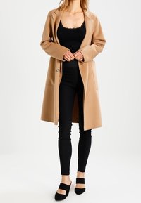 Kvinna klädd i en kamelfärgad kappa över en svart topp med spetskanter, svarta byxor och svarta klackskor, stående mot en ljus, enfärgad bakgrund.