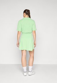 Camiseta polo verde combinada con una falda plisada a juego. La tela es suave con un ligero brillo. Se lleva con zapatillas blancas y calcetines.