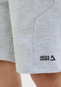 Pantaloni corti in cotone grigio con tasca laterale e logo nero "JACK & JONES" con design triangolare sull'orlo. Texture morbida e vestibilità casual.