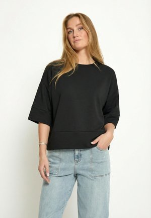 Mujer con cabello largo y rubio, vistiendo una blusa negra de manga corta y suelta, y jeans azul claro, de pie frente a un fondo claro y liso.