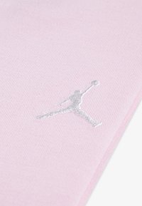 Jasnoróżowy materiał o miękkiej teksturze z srebrnym haftowanym logo Jumpman w pozycji w powietrzu. Proste i nowoczesne detale projektu.