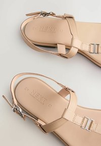 Beige leather sandals with ankle straps, featuring silver metal buckles and embossed brand name on the footbed. Flat sole design.