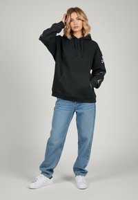 Hoodie nera con tasca frontale, cordino regolabile e dettagli ricamati sulle maniche. Abbinato a jeans azzurri chiari e sneakers bianche.