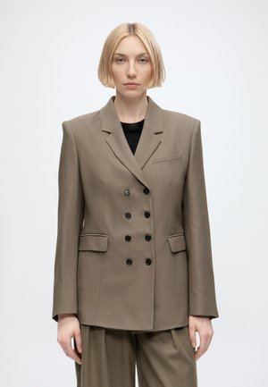 Vrouw met kort blond haar die een bruine double-breasted blazer en bijpassende wijde broek draagt tegen een egaal witte achtergrond.