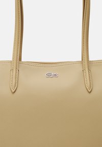 Sac fourre-tout beige en matériau texturé avec deux longues poignées. Présente un petit logo brodé dans des couleurs contrastées.
