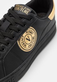 Svart lädersneaker med strukturerad yta, guld-accenterad logotyp-patch och svarta snörningar. Utrustad med gummisula och rund tådesign.