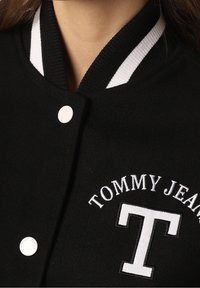 Černá univerzitní bunda se bílým pruhovaným límcem, zapínáním na knoflíky a vyšitým logem "TOMMY JEANS" s výrazným designem písmena "T".