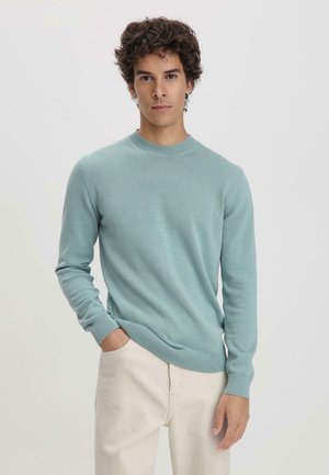 Hellblaues Strickpullover mit Rundhalskragen und langen Ärmeln. Merkmale sind gerippte Bündchen und Saum, kombiniert mit hellen Hosen.