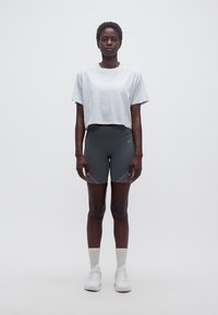 Γυναίκα που στέκεται στραμμένη προς τα εμπρός, φοράει λευκό κοντό μπλουζάκι, σκούρα γκρι ποδηλατικά σορτς, λευκές κάλτσες και λευκά αθλητικά παπούτσια πάνω σε απλό φόντο.