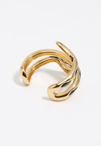 Pulsera de brazalete de metal dorado con un diseño abierto que presenta elementos curvados y ondulados, y acentos recortados, reflejando la luz con un acabado brillante.