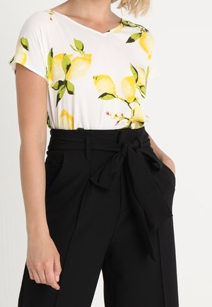 Kobieta ubrana w biały krótki top z żółtym wzorem cytryn, włożony do czarnych spodni z wysokim stanem związywanych paskiem w talii i kieszeniami.