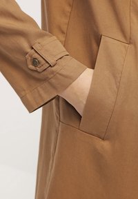 Main insérée dans la poche latérale d'un vêtement marron à manches longues avec détail de poignet boutonné.