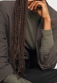 Capelli intrecciati che cadono su un blazer scuro e oversize con apertura frontale, sovrapposto a un top verde oliva testurizzato e pantaloni neri aderenti.