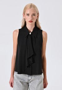 Modella che indossa un top nero senza maniche con un nodo e un bottone decorativo, abbinato a jeans in denim chiaro, su uno sfondo semplice.