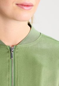 Veste zippée verte avec un col côtelé et une finition texturée. Présente une fermeture éclair argentée à l'avant, mettant en valeur un design minimaliste.