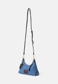 Blå denimväska med triangulär form, med svarta remmar, guldtoni detaljer och en röd och svart logopatch på framsidan.