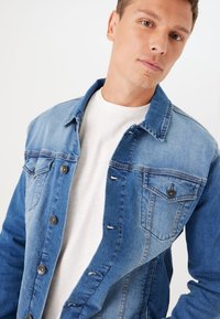 Chaqueta de mezclilla en diferentes tonos de azul con un cierre de botones en la parte delantera, bolsillos en el pecho y un cuello clásico, presenta un acabado ligeramente texturizado.