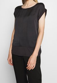 Dunkelgraue Satinbluse mit kurzen, umgeschlagenen Ärmeln und einem leicht verlängerten Saum in der Taille. Glatte Textur mit einem dezenten Glanz.