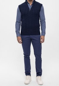 Námořnická pletená vesta s kabelovým vzorem, spárovaná s modrou košilí na knoflíky a námořnickými kalhotami. Bílá sneakers dodávají outfitu kompletní vzhled.