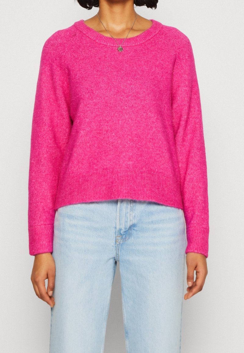Personne portant un pull rose vif à manches longues et un jean taille haute bleu clair, debout devant un fond uni.