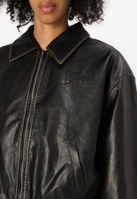 Svart läderjacka med krage, dragkedjestängning och en subtil broderad "Guess"-logotyp på bröstet. Texturerad yta synlig.