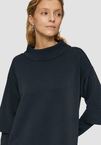 Navyblauwe oversized top met een hoge kraag, verlaagde schouders en een zachte textuur. Heeft lange mouwen en een relaxte pasvorm.