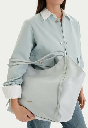 Mujer con chaqueta azul claro y vaqueros sosteniendo un gran bolso tote azul pálido con el logo dorado "JENNY".