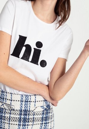 Person iført en hvid T-shirt med stor sort "hi." tekst og en højtaljet blå og hvid ternet nederdel, armen bøjet ved albuen.