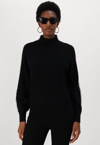 Pulover negru din tricot cu guler înalt, formă ajustată și accente albe pe mâneci. Model purtând ochelari de soare negri supradimensionați.