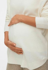 Un pull blanc en tricot superposé sur un vêtement beige, avec une main reposant sur un ventre arrondi, mettant en valeur des textures douces et une coupe décontractée.